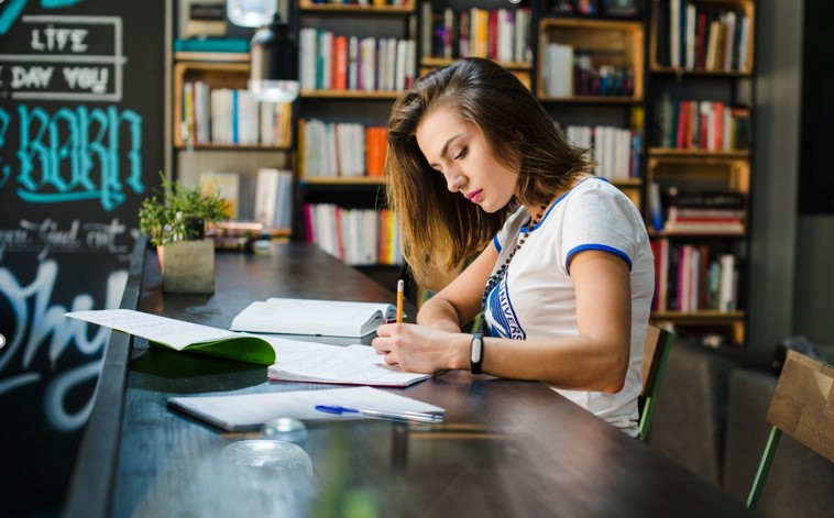 A young woman studies intently at a library table, writing in a notebook. Behind her are full bookshelves and modern decor, creating a focused atmosphere. Montlakepublishers